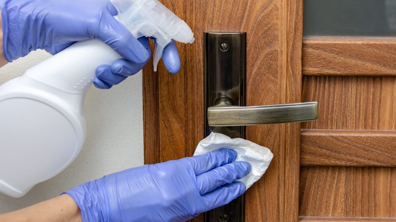 A cleaner in blue gloves spraying and disinfecting an office door handle